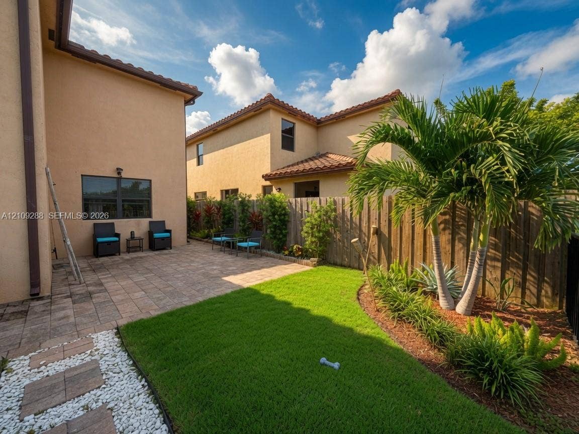 Stucco exterior with a paved stone patio, manicured lawn, wood privacy fencing, and palm trees