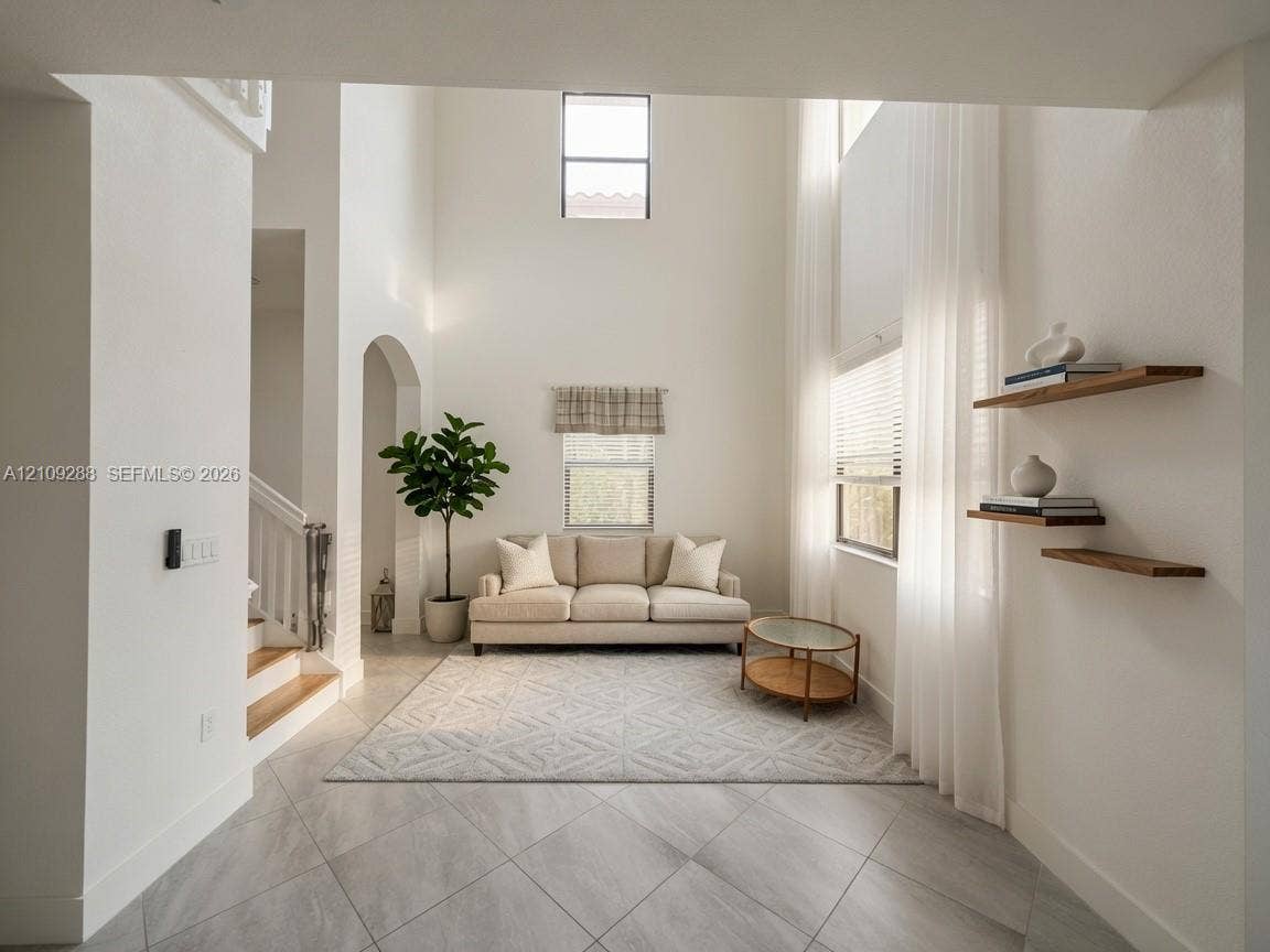 Double-height ceilings featuring natural lighting from multiple window levels, neutral stone-finish tile flooring, arched architectural detailing, and wall-mounted wood-finish floating shelves