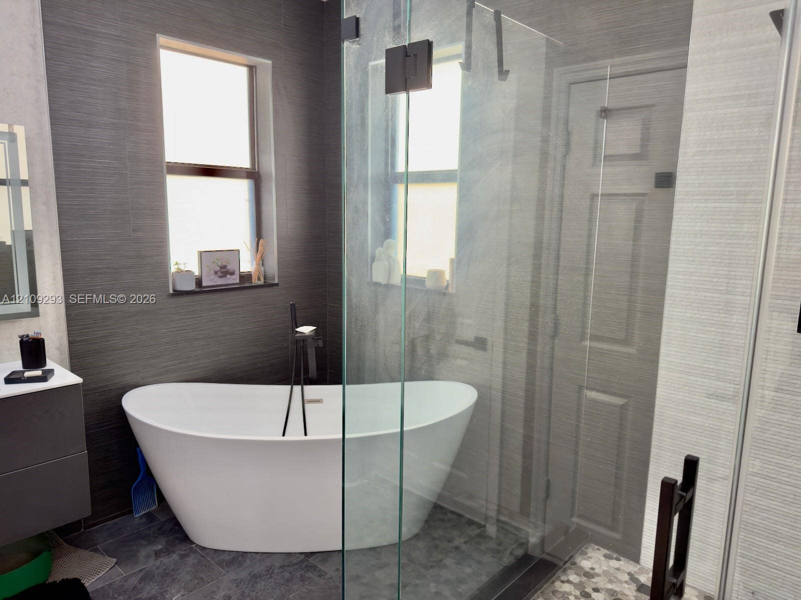 8880 NW 115th Court, Doral, FL 33178 Freestanding white soaking tub with matte black floor-mounted faucet, walk-in glass-enclosed shower, dark textured wall tiling, and floating vanity