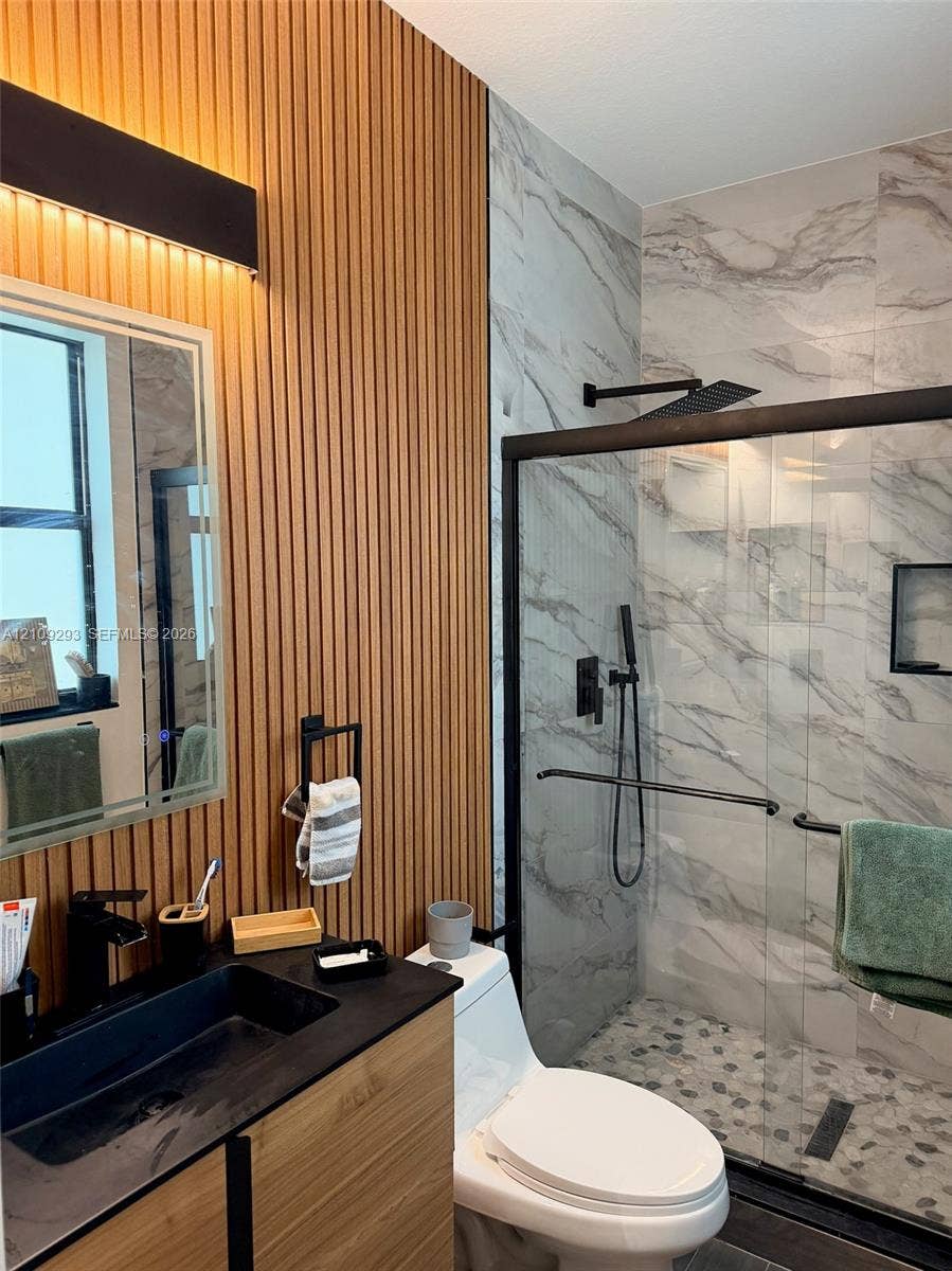 8880 NW 115th Court, Doral, FL 33178 Bathroom featuring a wood-slat accent wall, a floating vanity with a rectangular vessel sink, and a walk-in shower with stone-tile flooring, floor-to-ceiling wall tile, and matte black fixtures