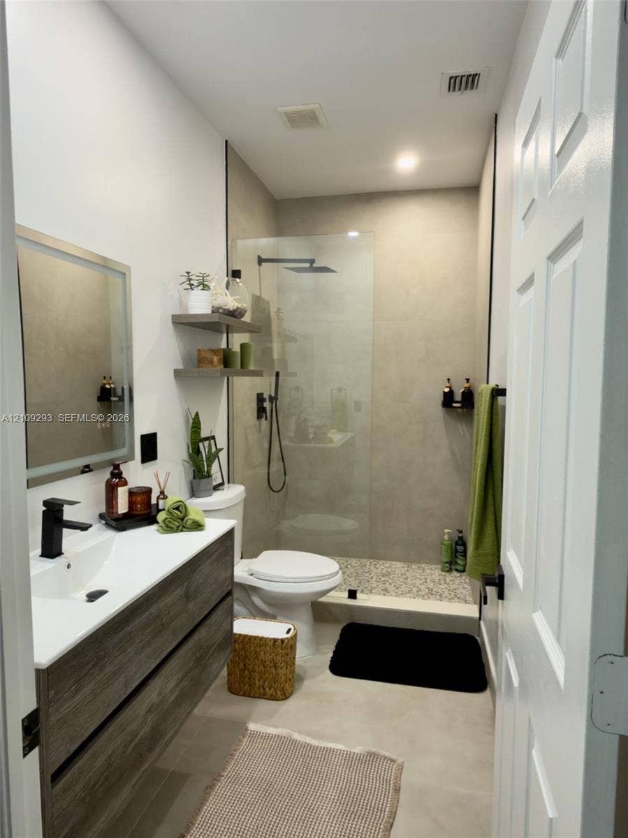8880 NW 115th Court, Doral, FL 33178 Floating wood-finish vanity with a white integrated sink, matte black faucet, illuminated mirror, walk-in shower with matte black rainfall fixture, and neutral large-format tile flooring