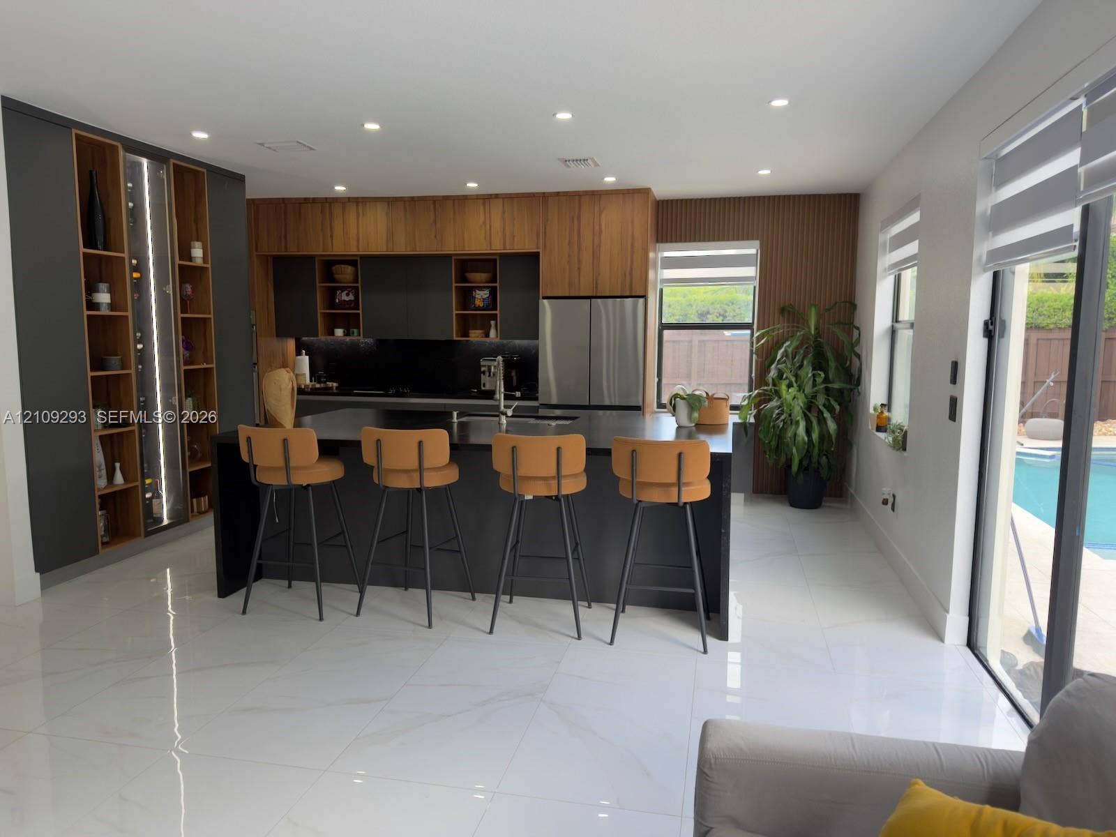 8880 NW 115th Court, Doral, FL 33178 Kitchen featuring custom cabinetry with wood-finish and matte-black accents, central island with dark stone countertop, integrated floor-to-ceiling shelving, recessed lighting, and large-format tile flooring