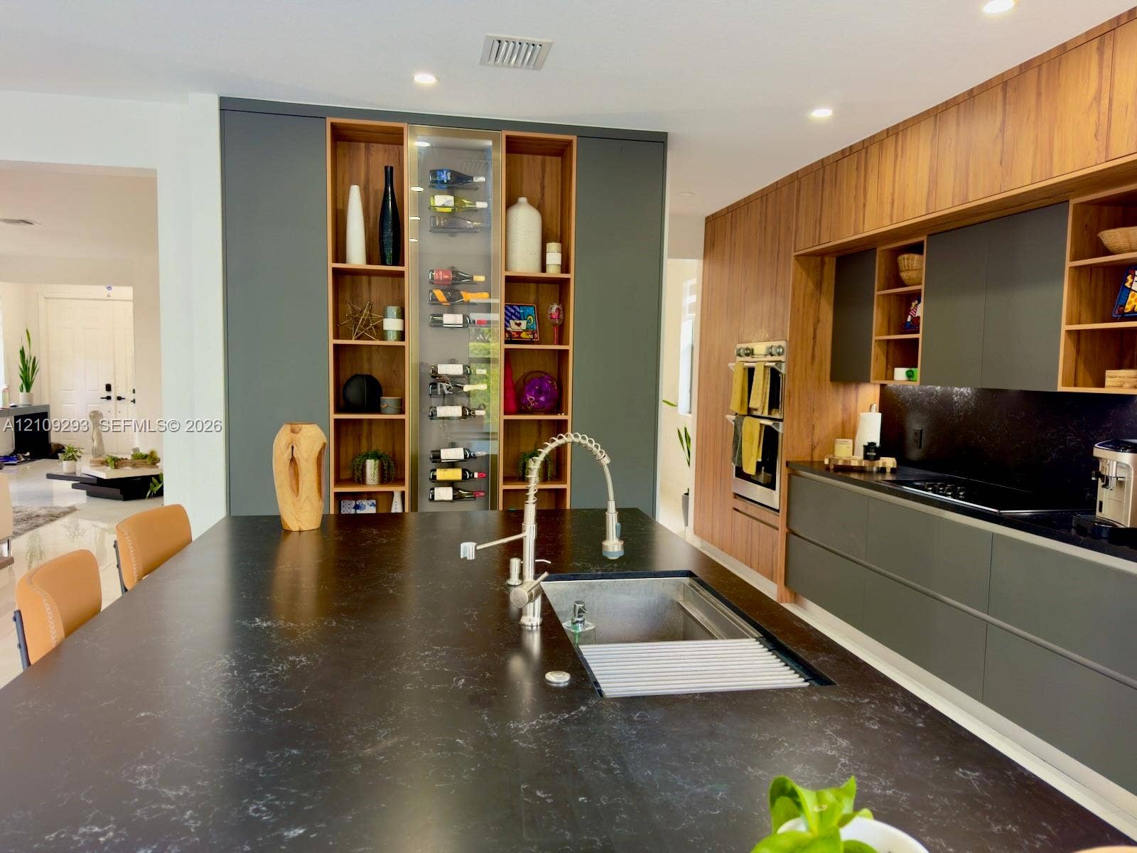 8880 NW 115th Court, Doral, FL 33178 Modern kitchen featuring custom cabinetry in dark gray and wood finishes, a large island with a dark solid surface countertop, built-in stainless steel double ovens, and a dedicated vertical wine storage display