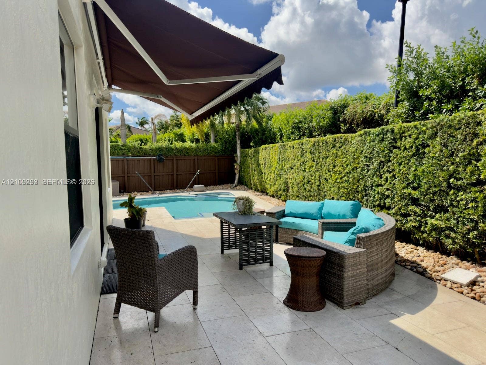 8880 NW 115th Court, Doral, FL 33178 Patio space featuring light-toned stone tile flooring, a retractable fabric awning, a swimming pool, and a perimeter privacy hedge