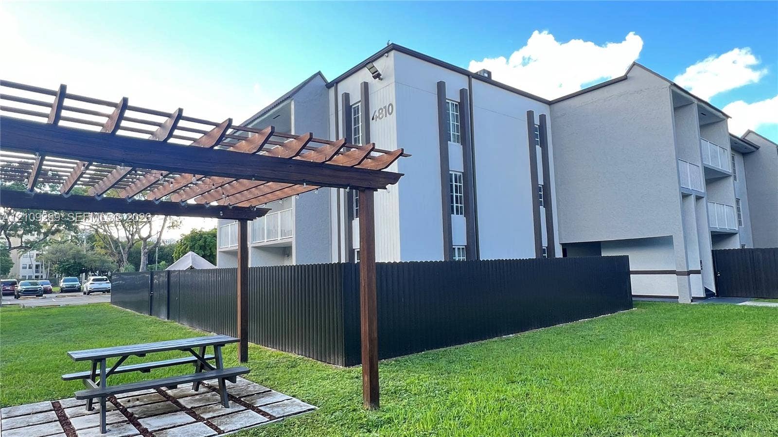 4810 NW 79 Avenue, #107, Doral, Doral, FL 33166 Exterior featuring a multi-story structure with a contemporary facade, wood-frame pergola, metal perimeter fencing, and landscaped grounds