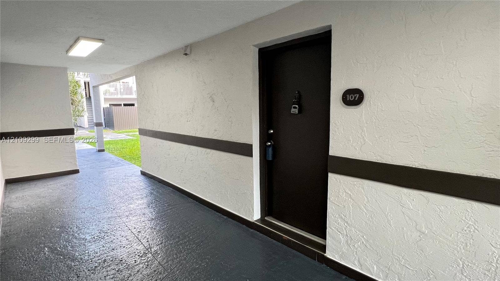 4810 NW 79 Avenue, #107, Doral, Doral, FL 33166 Covered exterior entryway featuring a dark-toned door with a circular unit number plaque, textured light-colored walls with horizontal trim, and painted concrete flooring