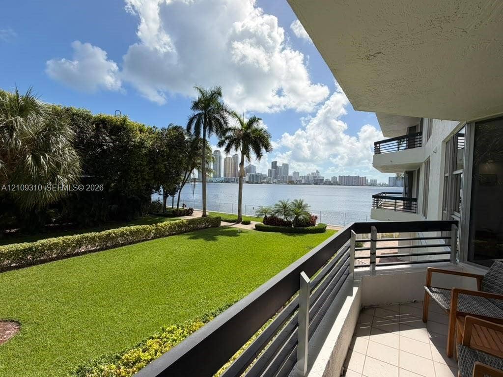 Covered balcony featuring tile flooring and horizontal metal railings, overlooking a landscaped lawn with mature palm trees and water frontage