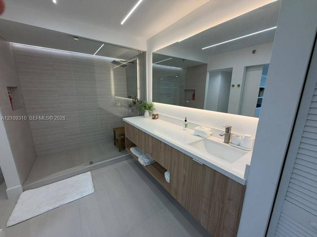 Floating wood-finish vanity with a white countertop and integrated sink, frameless glass walk-in shower with horizontal tile wall detailing, and a large-format LED-backlit mirror