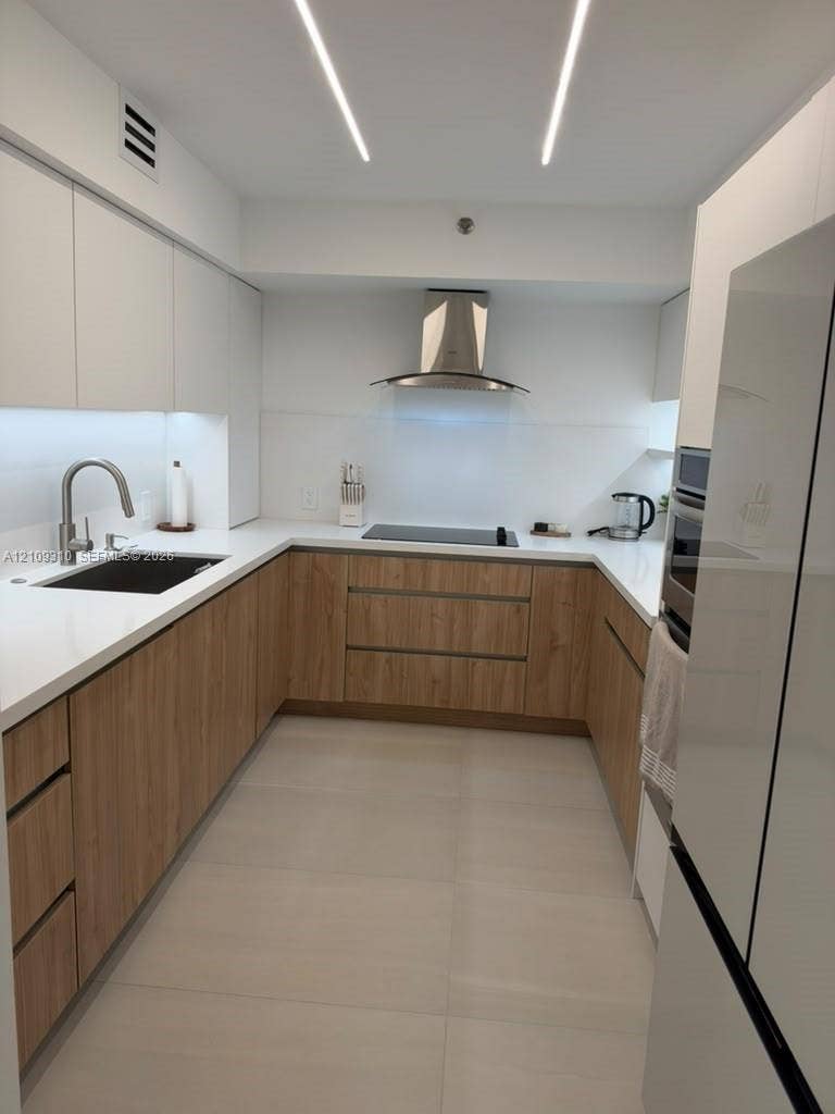 Modern kitchen featuring wood-finish cabinetry, solid-surface countertops, undermount sink with gooseneck faucet, stainless steel range hood, and linear recessed lighting