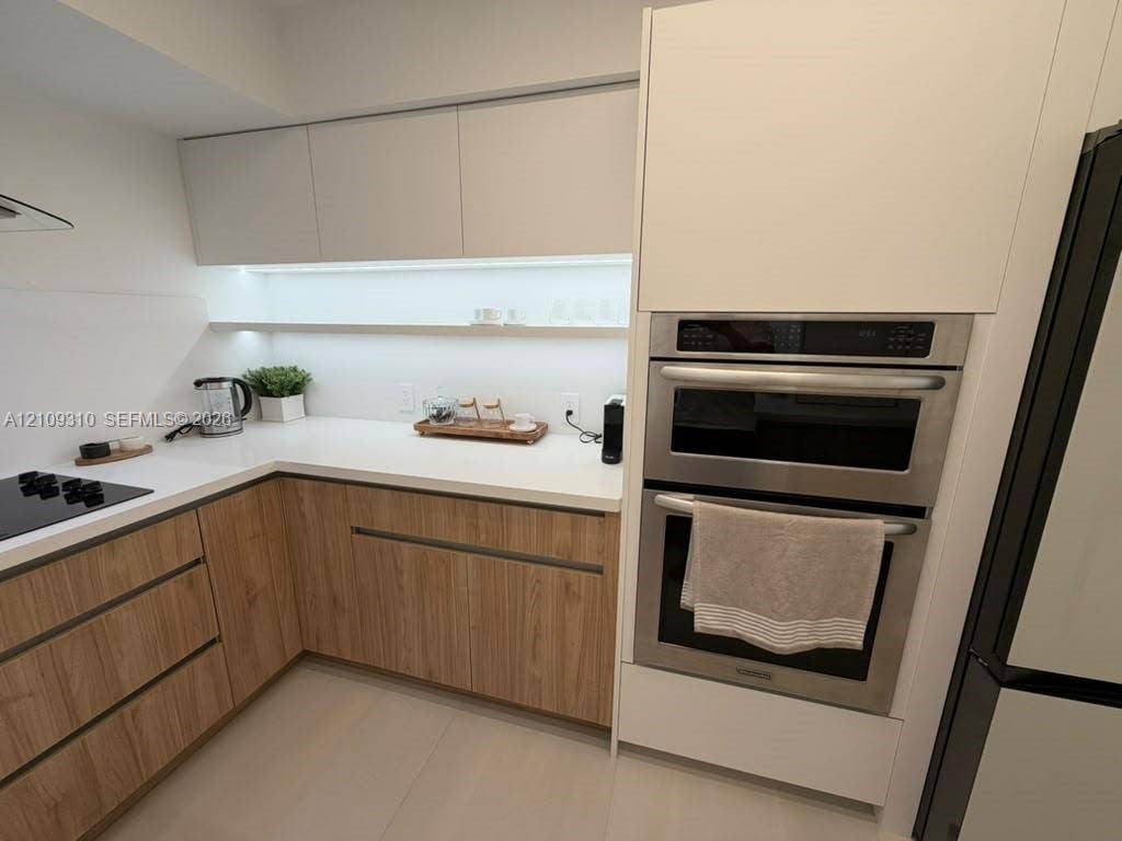 Kitchen featuring two-tone cabinetry with wood-finish lower units, white upper cabinets, white solid-surface countertops, integrated under-cabinet lighting, and a stainless steel wall oven
