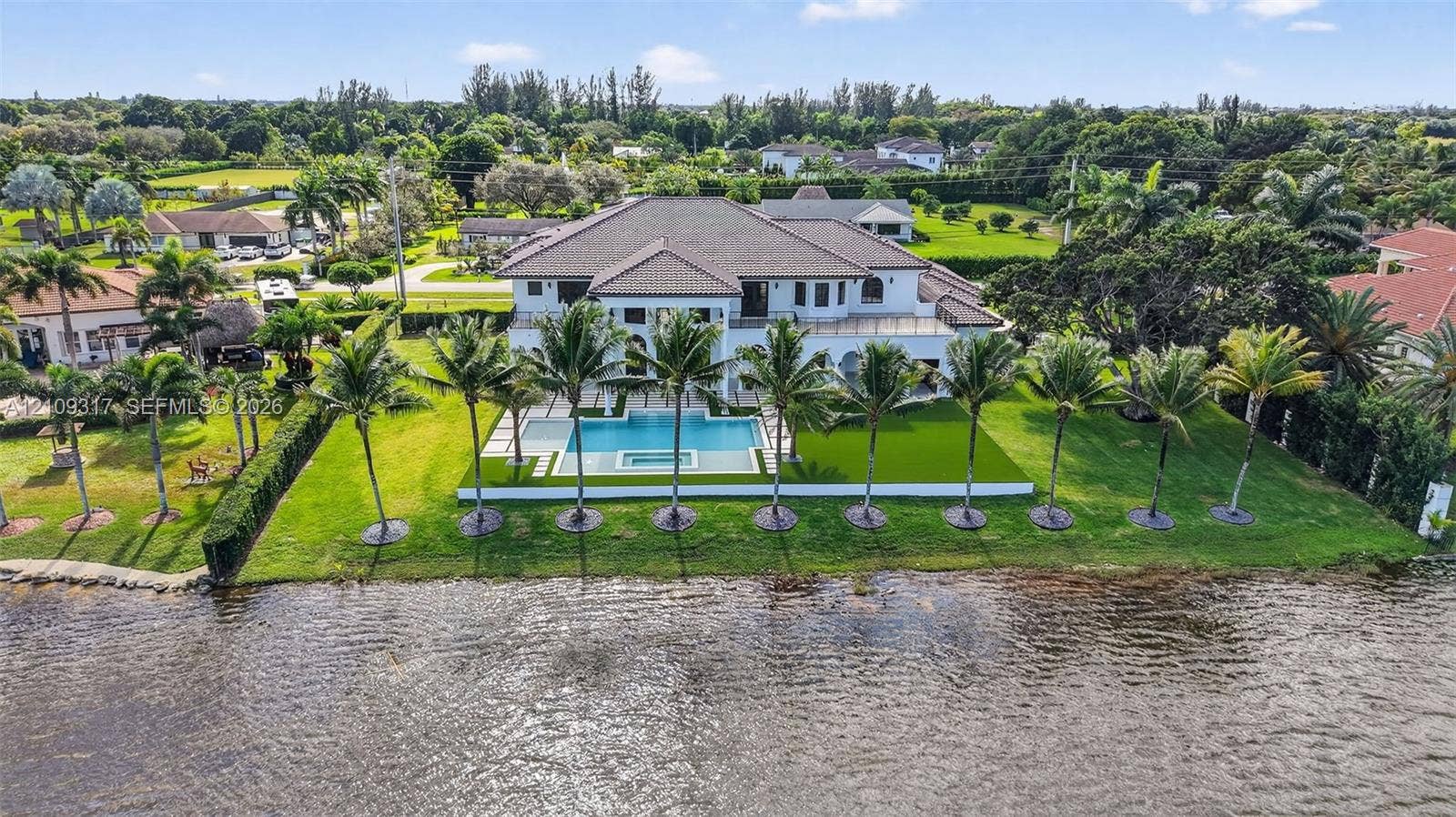 Waterfront estate featuring a multi-level roofline, swimming pool with integrated spa, paved terrace, and extensive manicured landscaping with mature palm trees