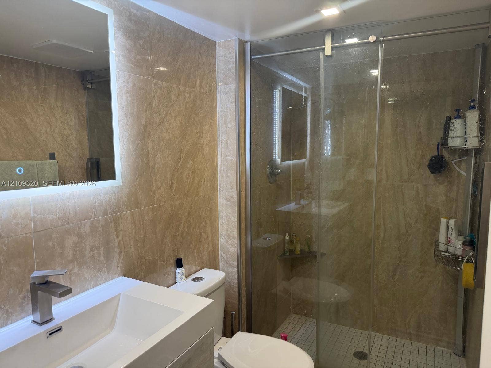 Full-height wall tiling featuring natural stone-patterned surfaces, a wall-mounted LED-backlit mirror, a white rectangular vessel sink with a single-handle chrome faucet, and a glass-enclosed shower with tiled flooring