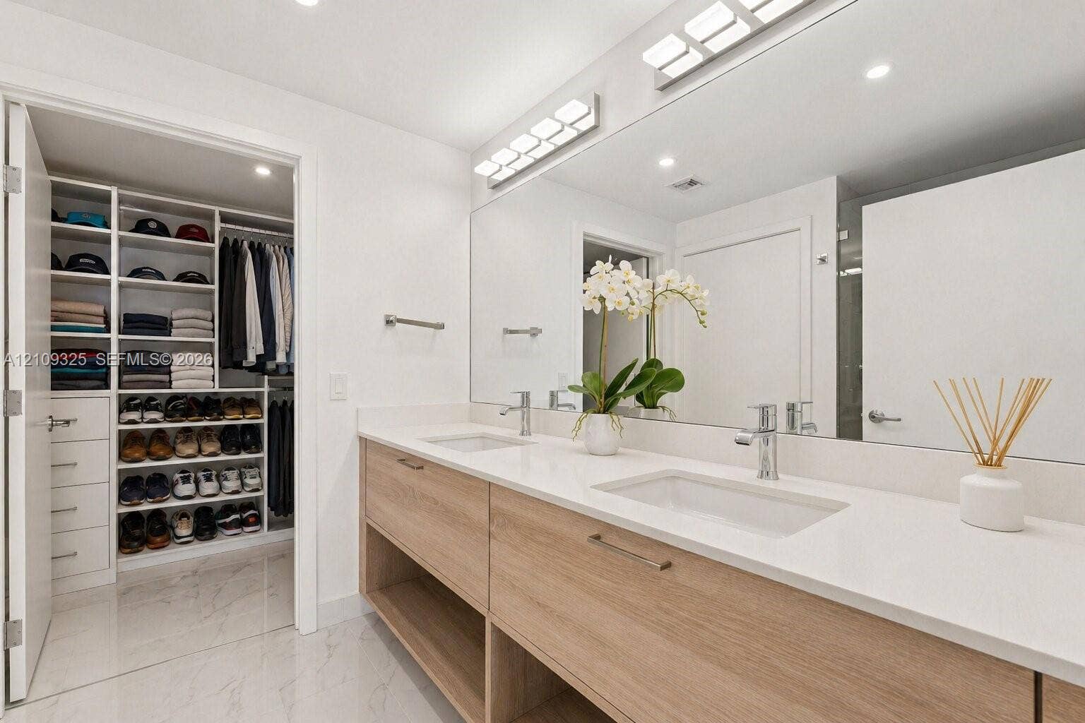 Dual-sink vanity with light wood-finish cabinetry, white solid-surface countertop, floor-to-ceiling walk-in closet with built-in shelving, and large-format tile flooring