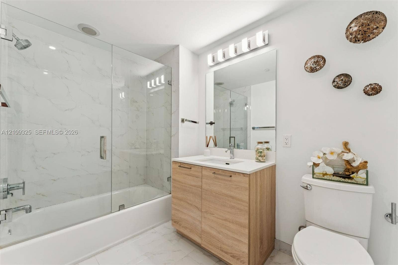 Bathroom featuring a glass-enclosed shower and tub combination with wall-to-wall surfacing, a vanity with wood-finish cabinetry, a white countertop with an integrated sink, and an expansive vanity mirror with overhead lighting