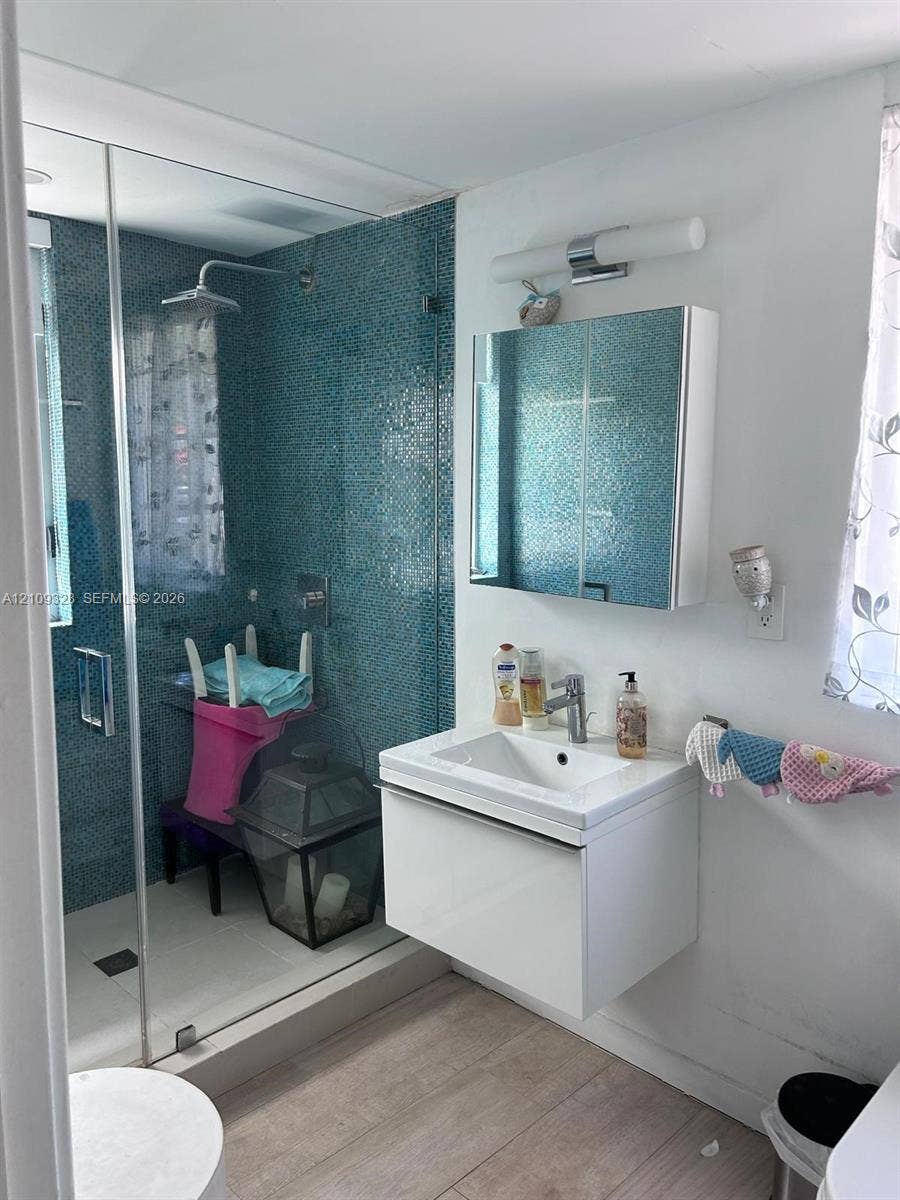 Floating white vanity with integrated basin, walk-in shower featuring blue mosaic tile surround, frameless glass enclosure, wall-mounted mirrored medicine cabinet, and wood-finish flooring