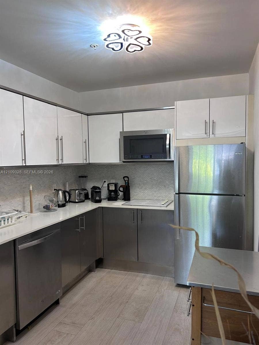 Kitchen featuring white upper cabinetry, stainless steel lower cabinetry, solid-surface countertops, textured tile backsplash, and stainless steel appliances