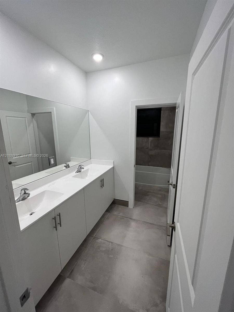 Dual-sink vanity with white cabinetry, sleek hardware, stone countertop, large wall-to-wall mirror, and oversized tile flooring