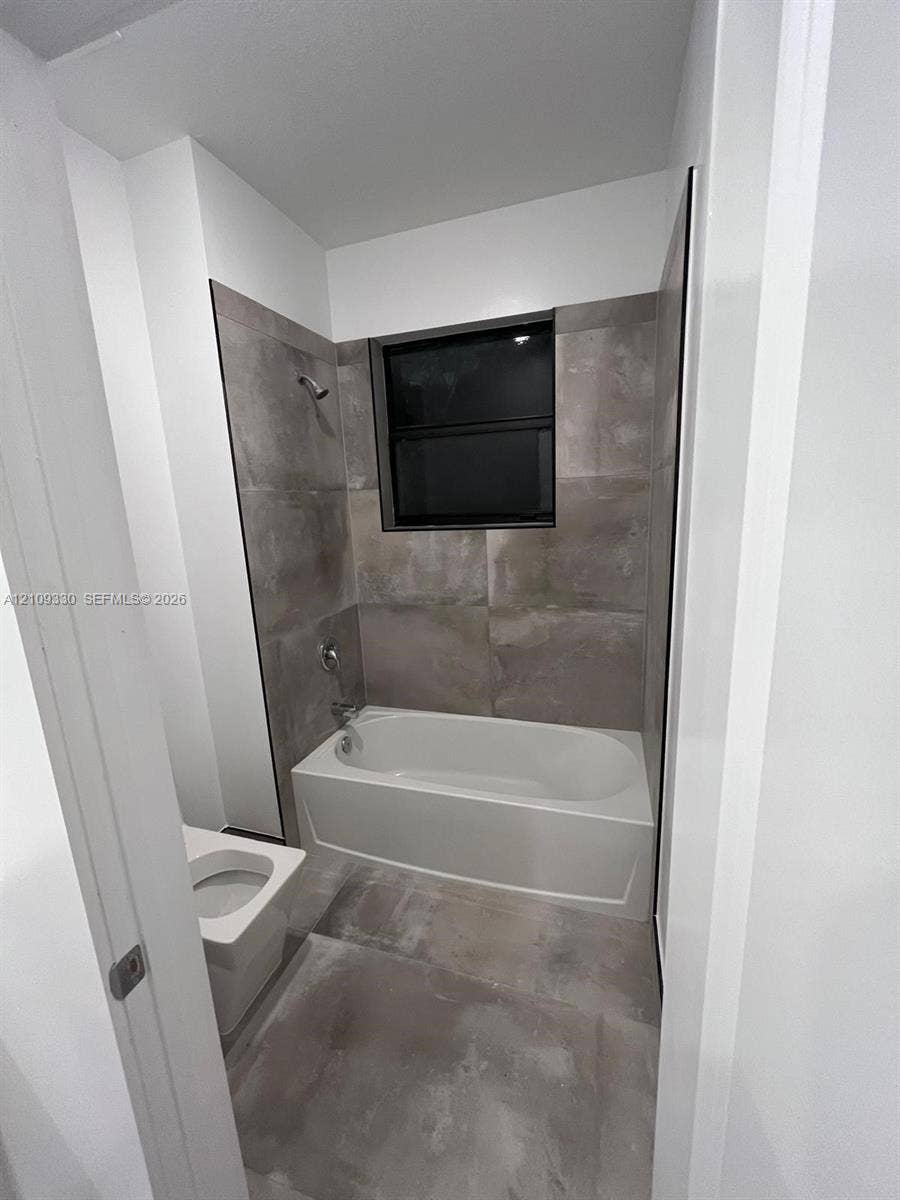 Full-height tiled walls surrounding a soaking tub with integrated shower hardware, large-format tile flooring, and a window with a black frame