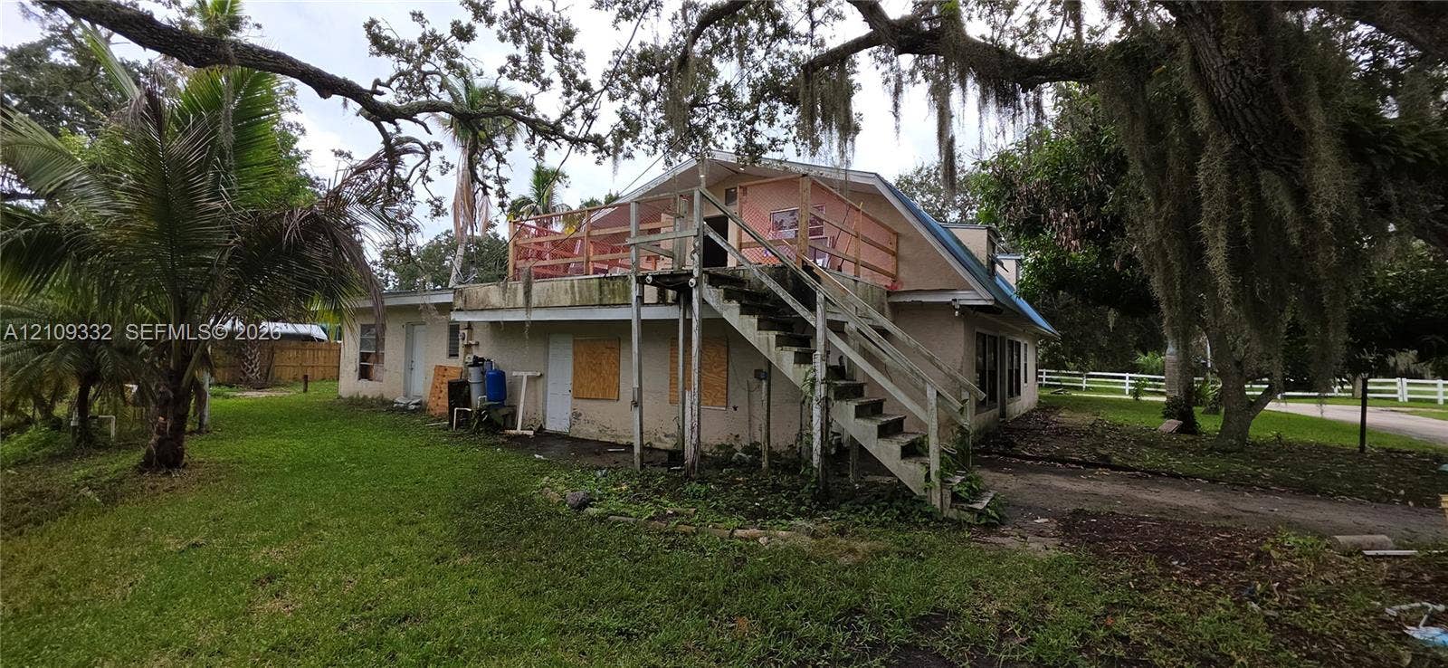 Two-story residential structure featuring an elevated wood deck, exterior staircase, and mature tree canopy on the grounds