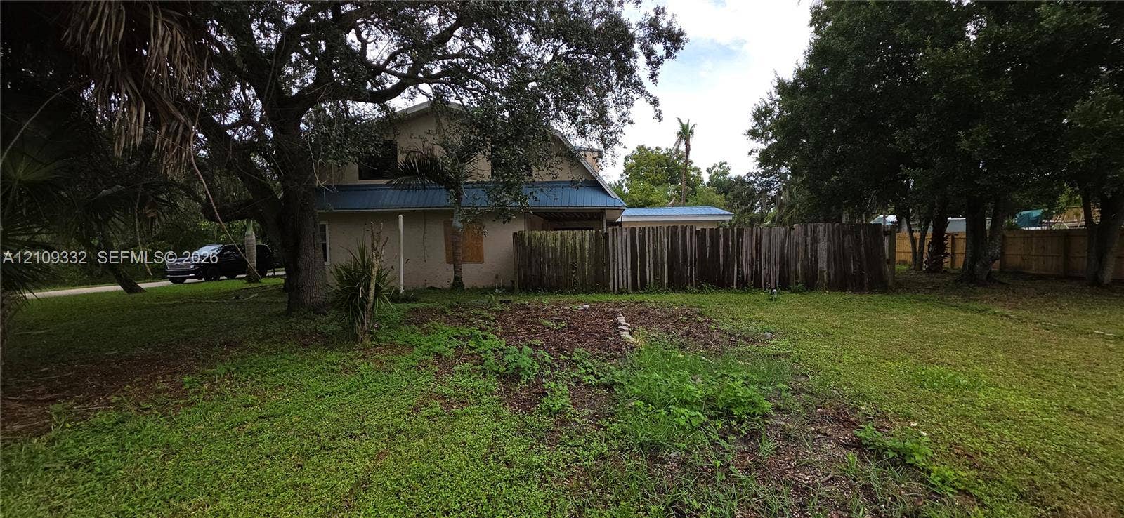 Exterior view featuring a multi-story residence with a blue metal roof, mature oak trees, and a wooden privacy fence
