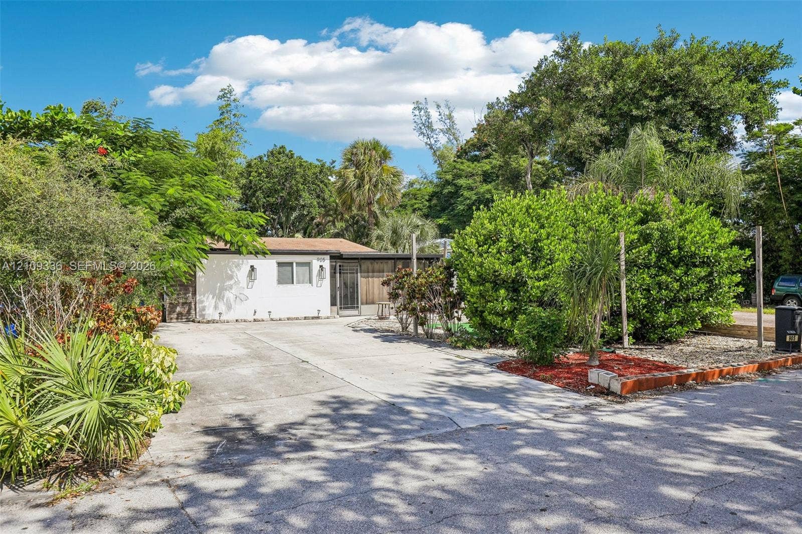Single-story exterior featuring a white stucco facade, concrete driveway, and established landscape including mature trees and shrubbery