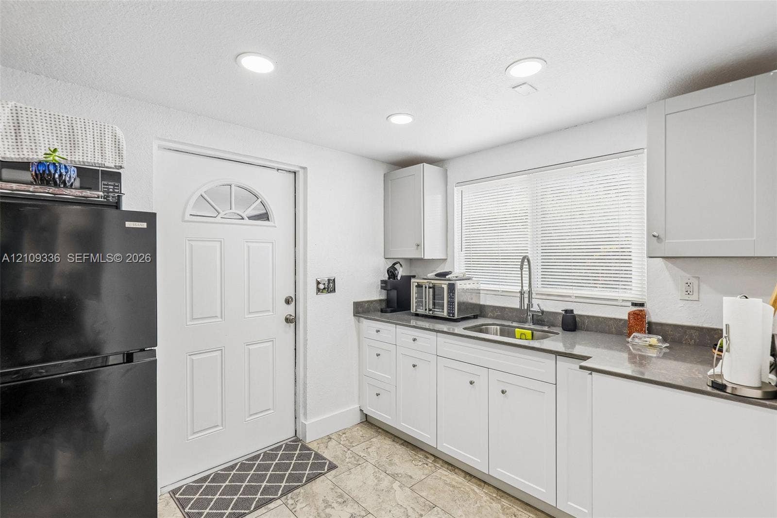 Kitchen featuring white Shaker-style cabinetry, dark stone-finish countertops, stainless steel undermount sink with arched pull-down faucet, tile flooring, and recessed ceiling lighting