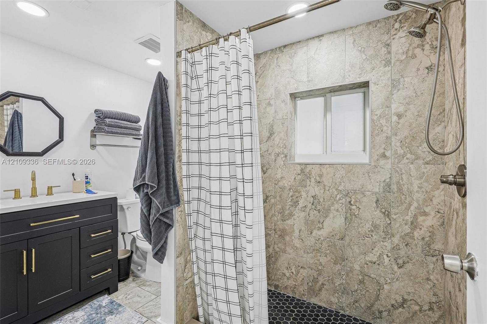 Bathroom featuring a dark-finish vanity with gold-tone hardware, stone-surround shower with a handheld fixture, hexagonal tile shower floor, and a framed wall mirror