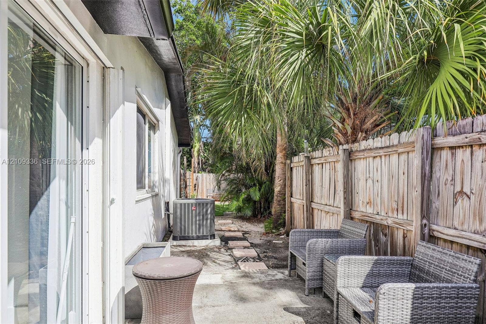 Side patio featuring concrete pavers, wood-panel fencing, mature palm trees, and sliding glass door access
