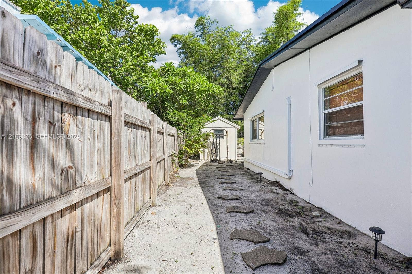 Side yard featuring a tall wood privacy fence, white stucco exterior siding, stone paver path, and a detached storage shed