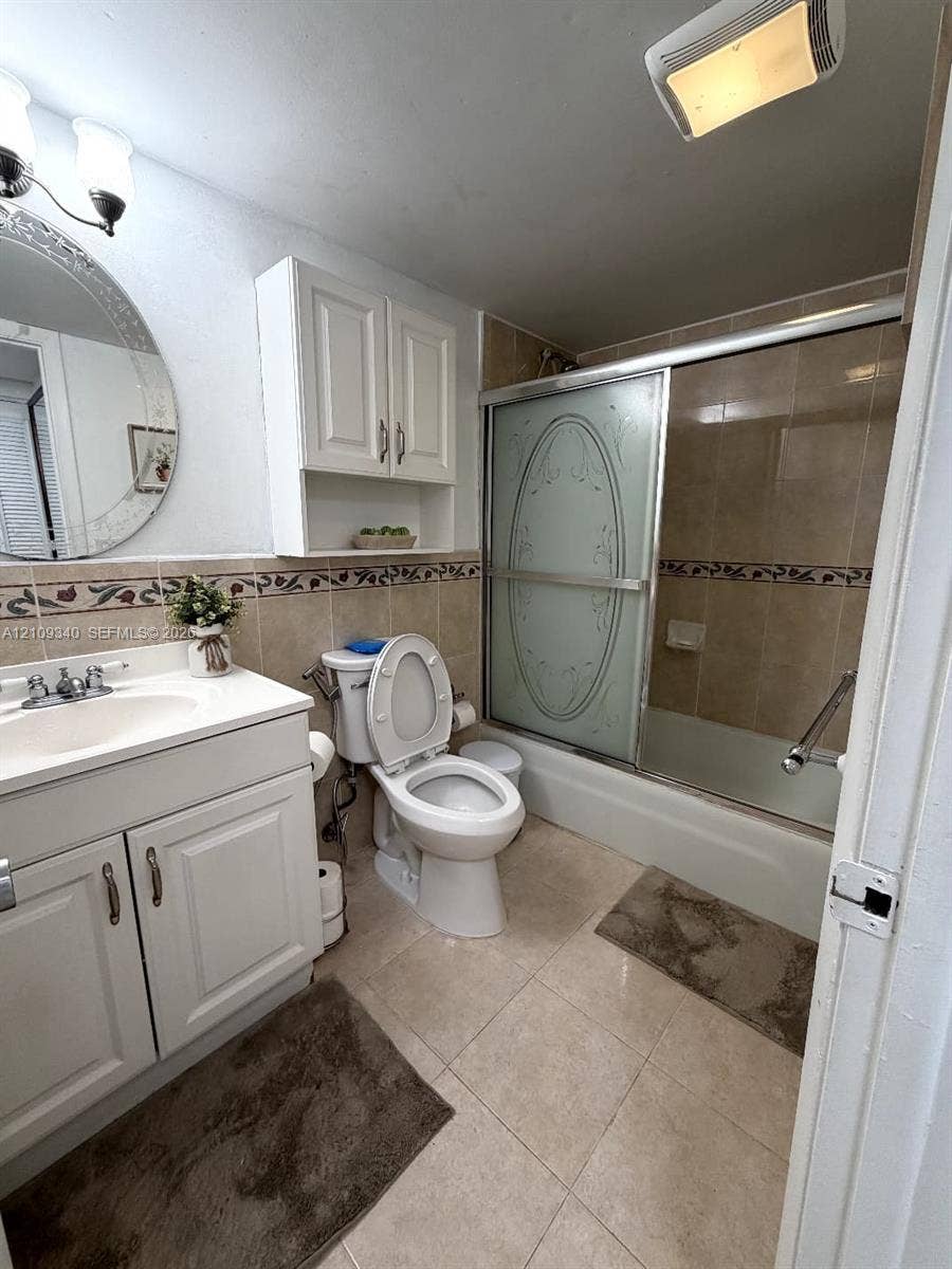 Bathroom featuring a white vanity cabinet with an integrated sink, wall-mounted storage cabinetry, a recessed bathtub with a decorative glass sliding door, and tile flooring