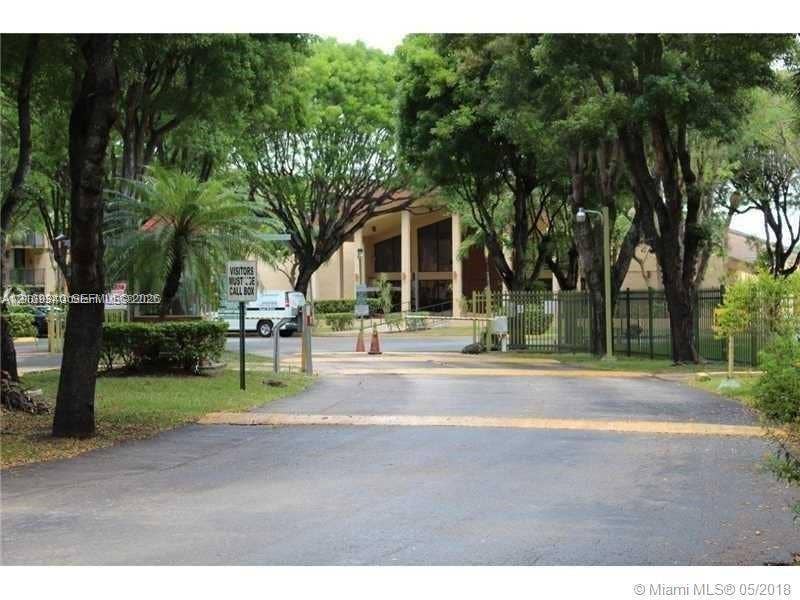 Paved entrance featuring mature canopy trees, metal perimeter fencing, and an gated access point
