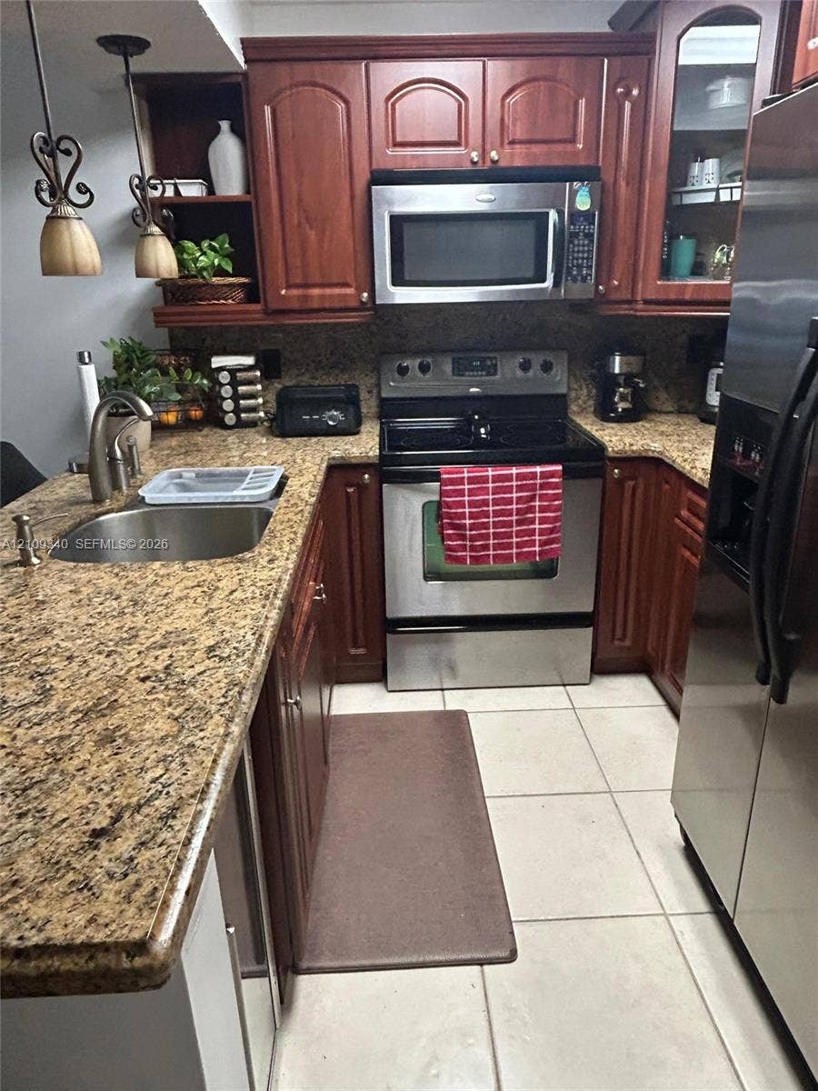 Kitchen featuring stained wood cabinetry, stone countertops, stainless steel appliances, ceramic tile flooring, and two pendant lights