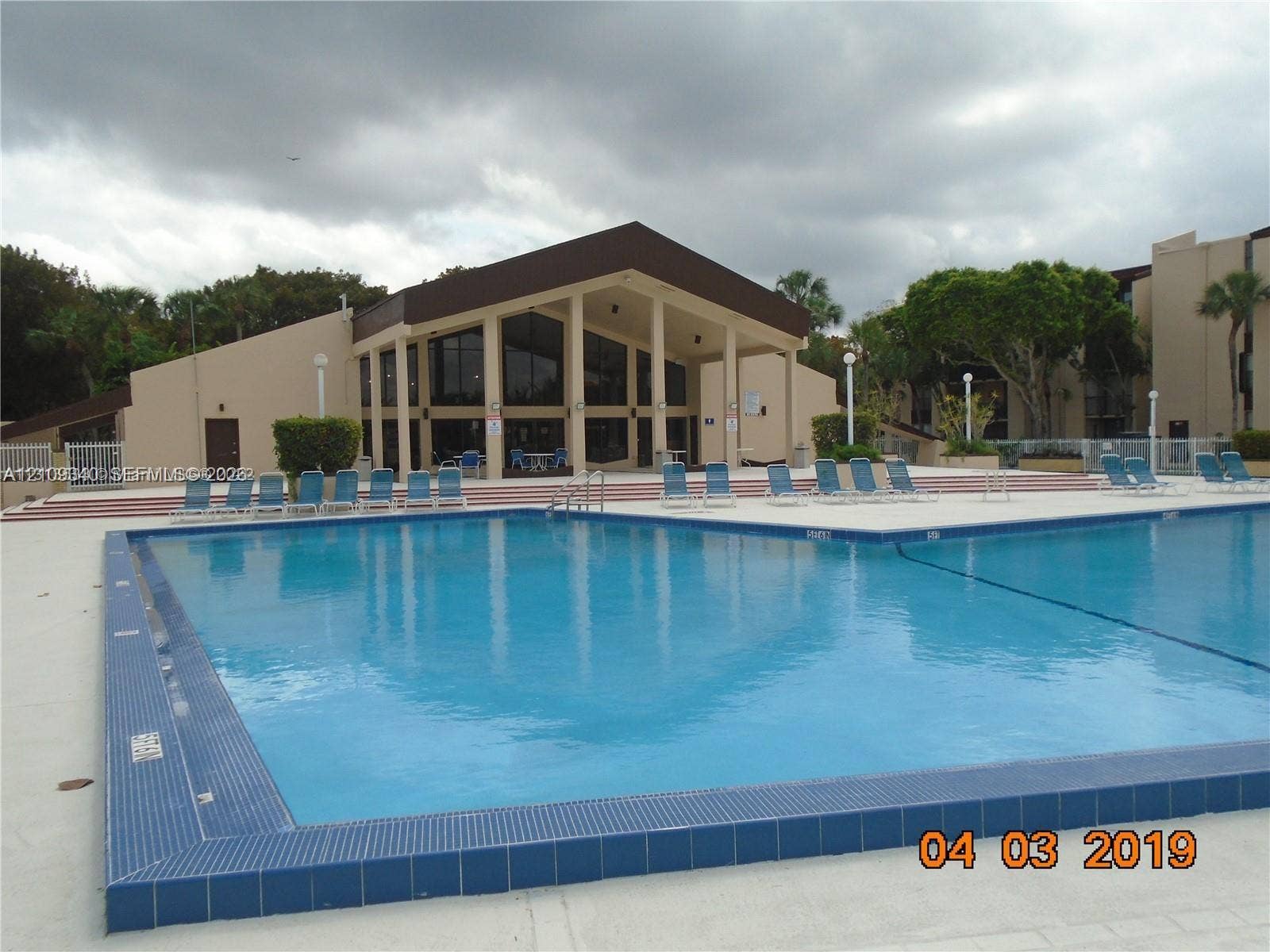 In-ground swimming pool with a tiled perimeter, expansive concrete deck, and multi-level structure featuring a gabled roof and floor-to-ceiling windows