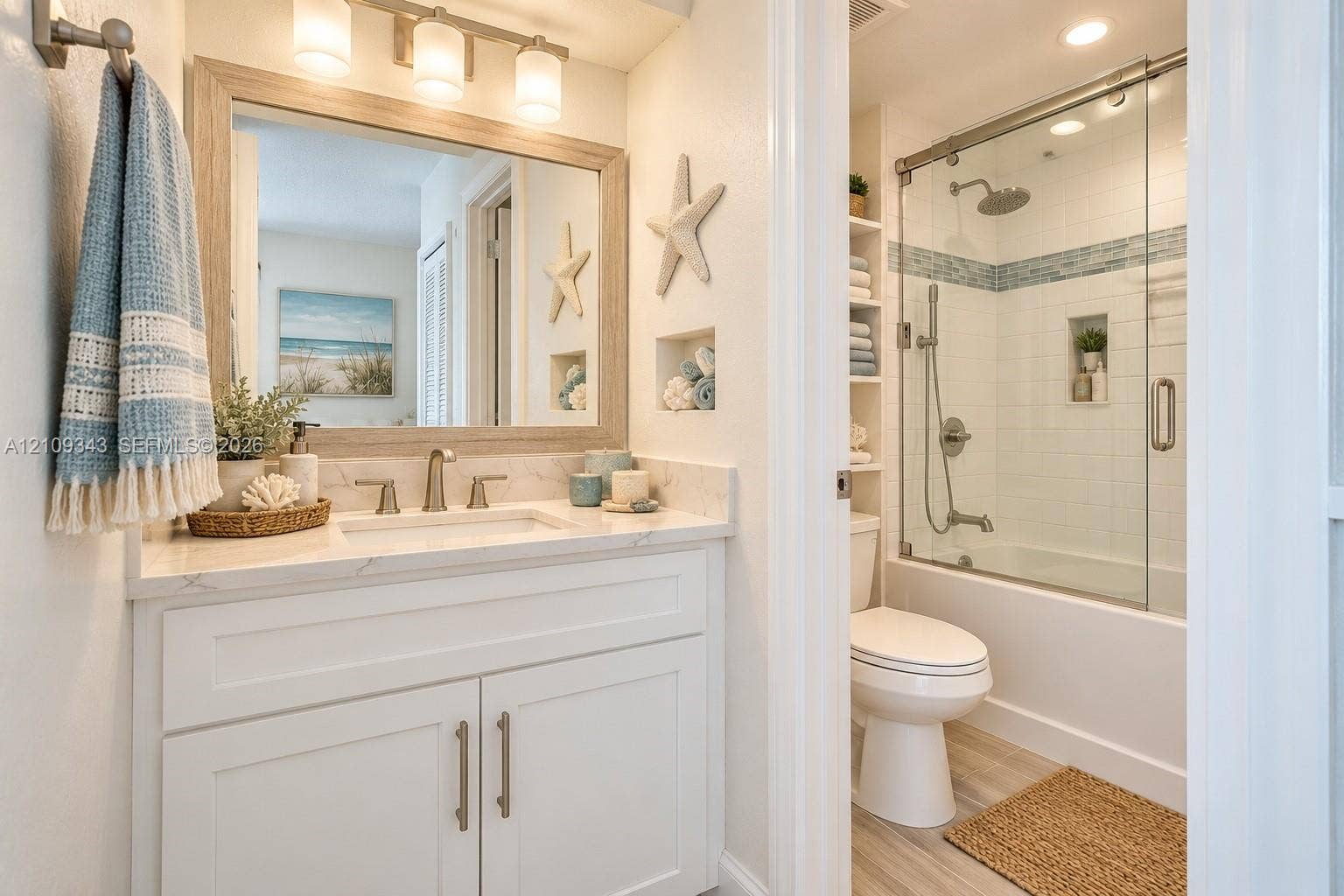 Bathroom featuring a white shaker-style vanity with stone-finish countertop, undermount sink, framed mirror with three-light fixture, built-in shelving, and a tub-shower combination with sliding glass doors and recessed wall niche