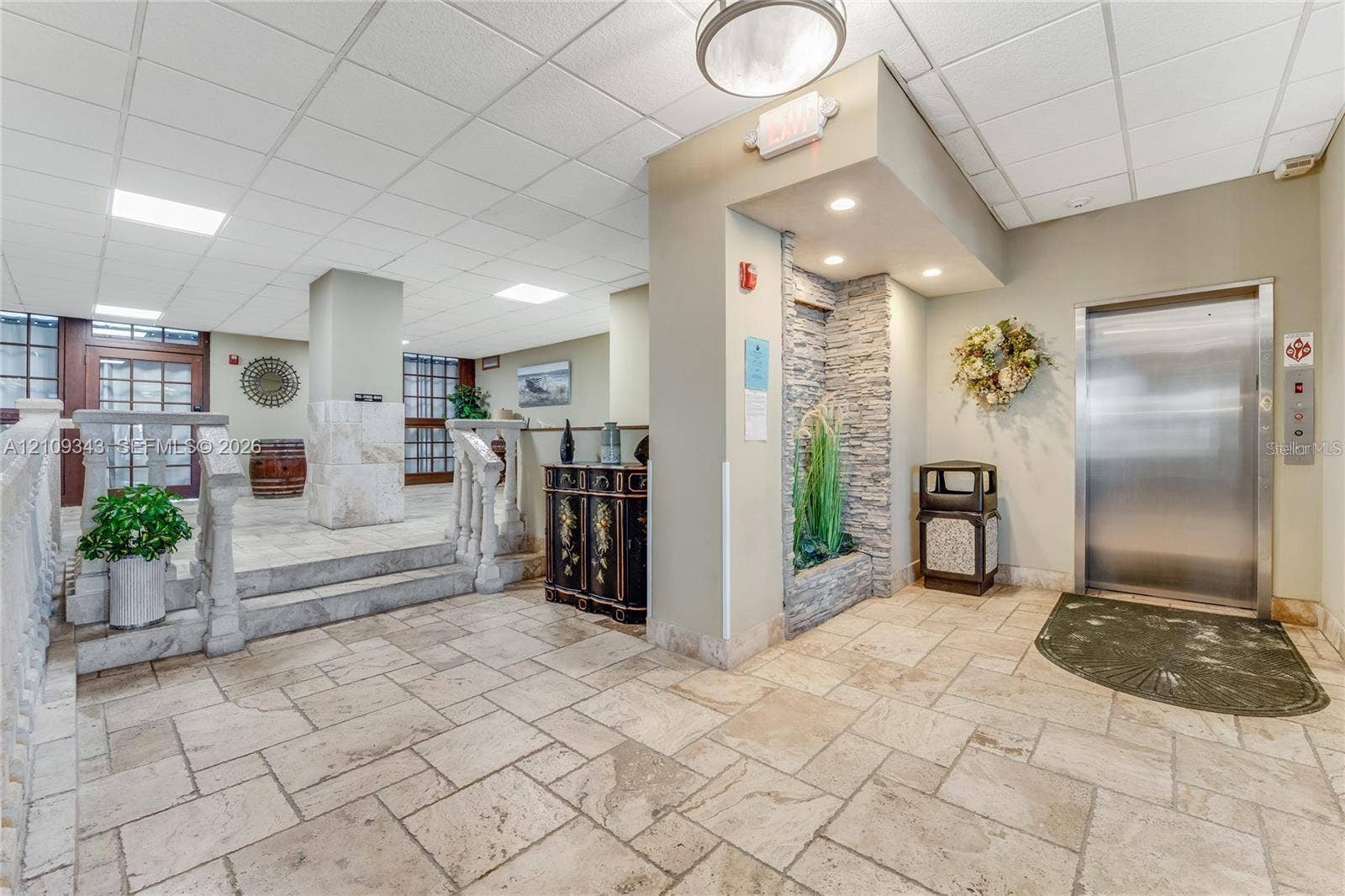 Lobby entrance featuring stone tile flooring, a decorative stone-clad accent wall with an integrated water feature, stainless steel elevator, and a balustrade staircase