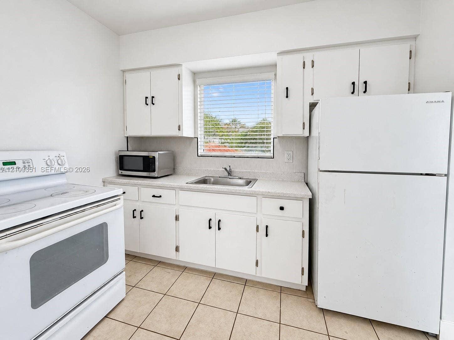 Kitchen featuring white cabinetry with black hardware, stone-composite countertops, stainless steel sink, tile flooring, and integrated white appliances