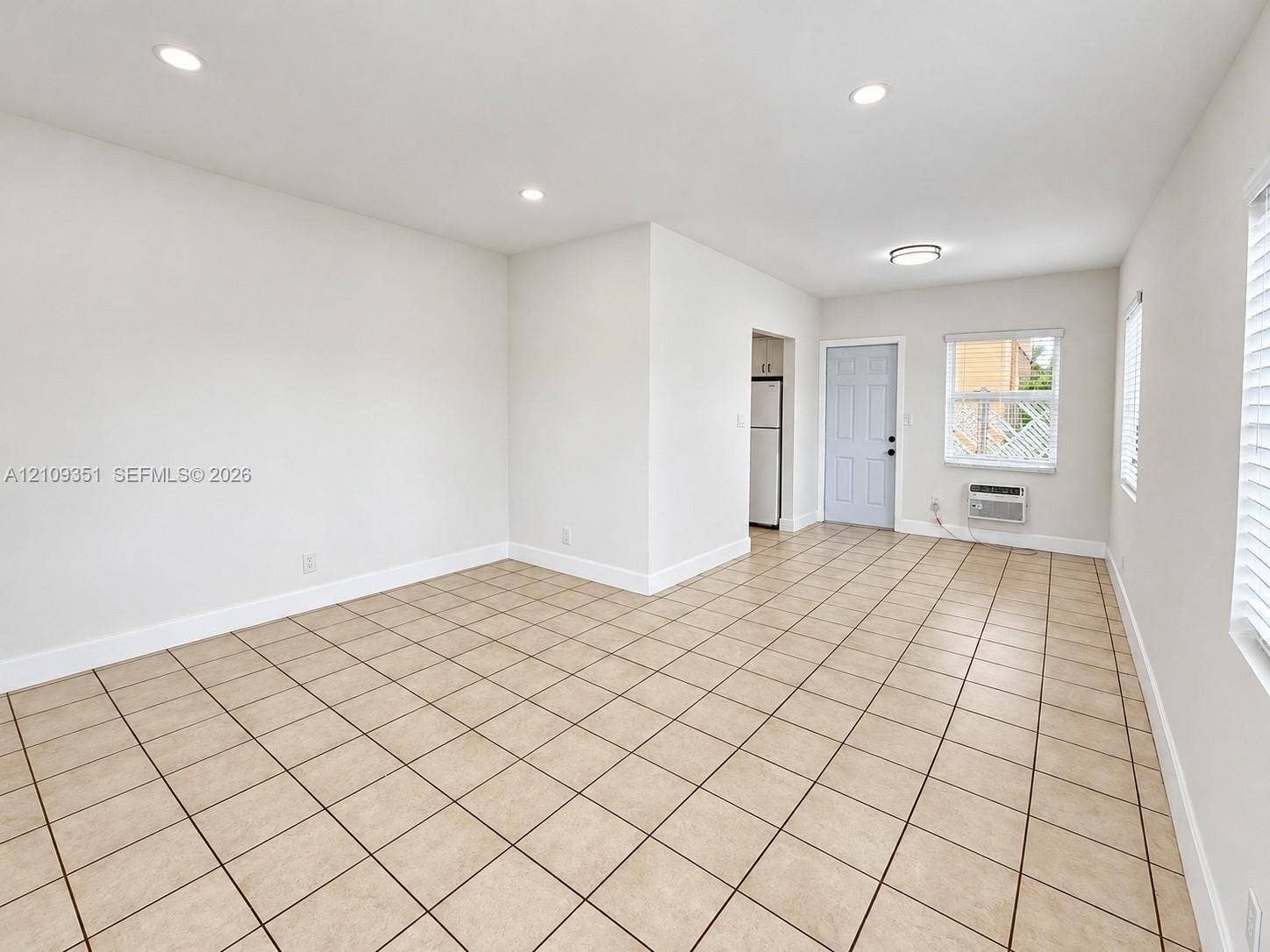 Neutral-toned tile flooring, recessed ceiling lighting, wall-mounted air conditioning unit, and large window with integrated blinds