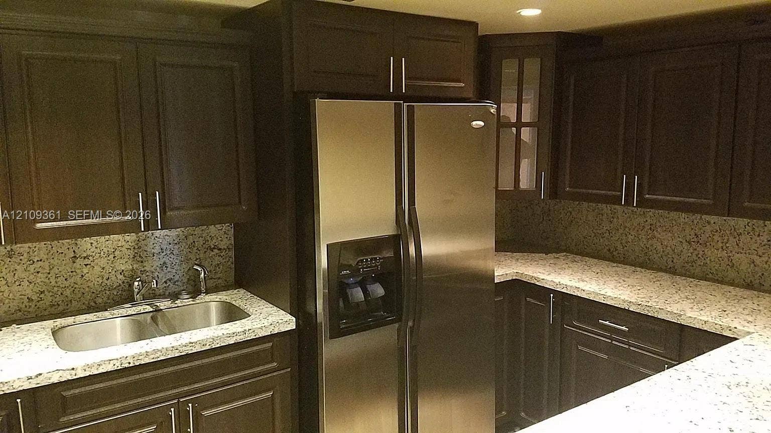 Dark wood cabinetry featuring brushed metal hardware, speckled stone countertops, stainless steel side-by-side refrigerator, under-mounted double-basin sink, and glass-paneled upper cabinet