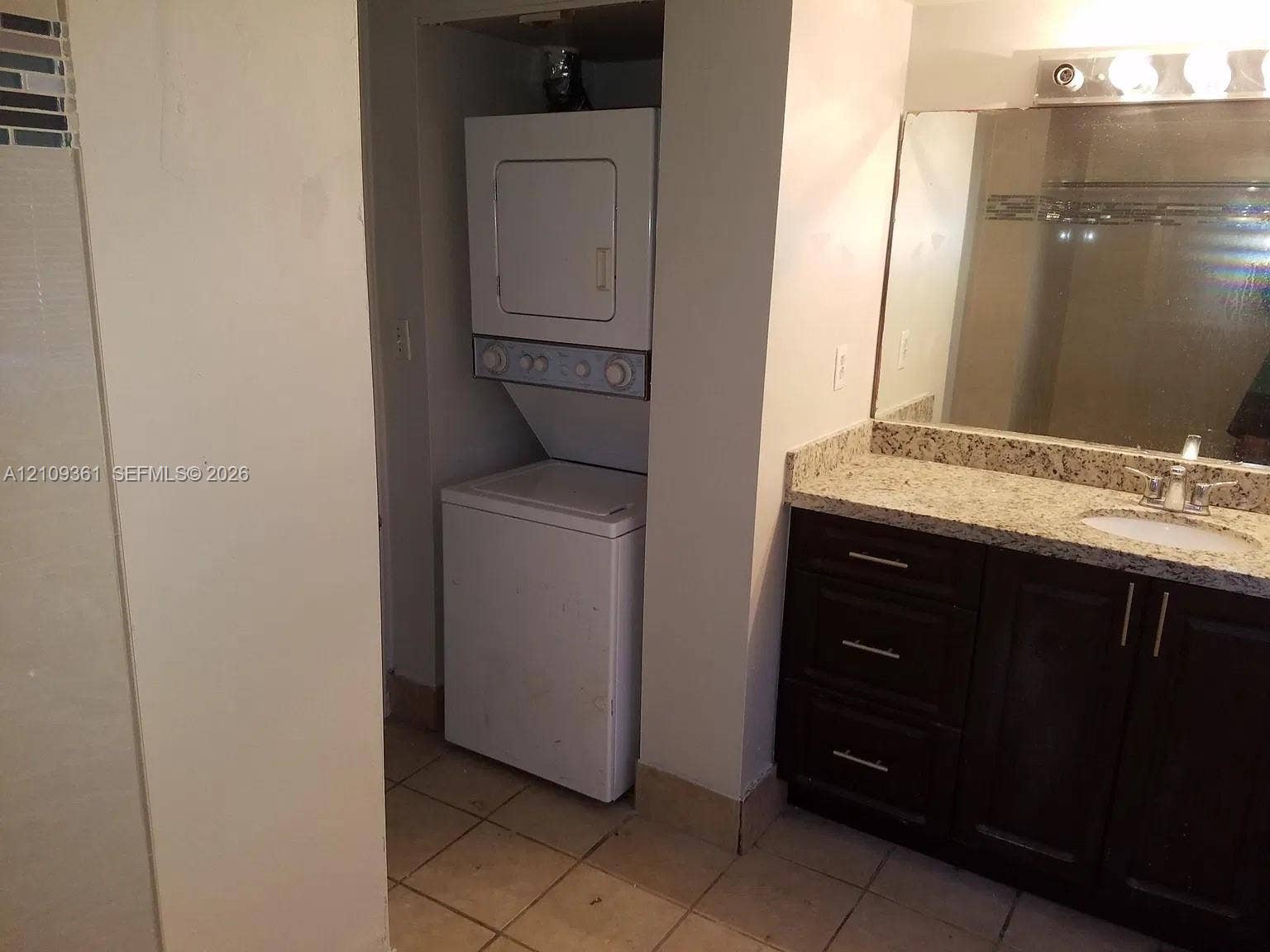 Stacked washer and dryer, vanity featuring stone-composite countertop, undermount sink, dark-finish cabinetry with metallic hardware, and full-width mirror