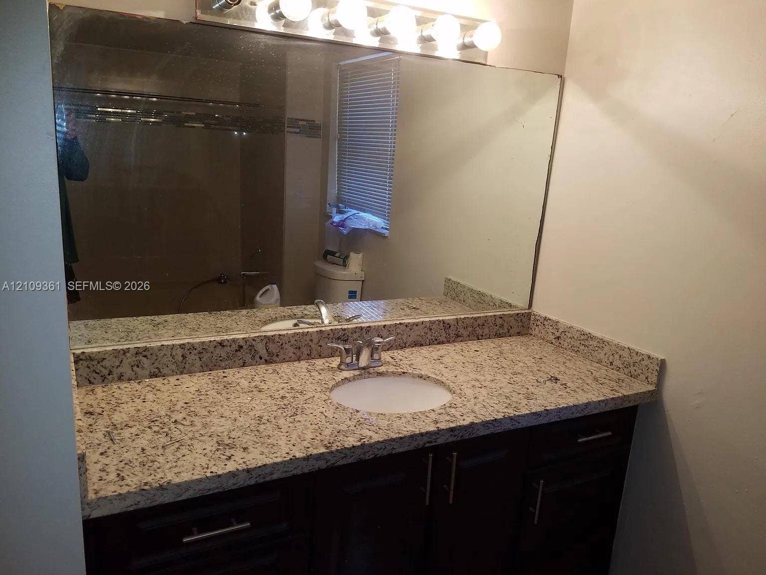 Vanity with stone-finish countertop, undermount sink, dual-handle faucet, dark-finish cabinetry, and full-width wall mirror with attached multi-bulb lighting fixture