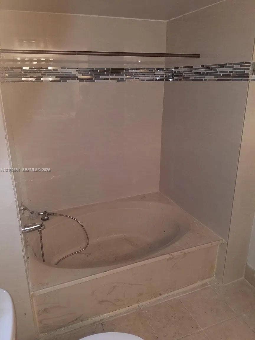 Built-in alcove soaking tub featuring a tiled surround, integrated stone-finish deck, mosaic glass accent tiling, and a chrome hand-held shower fixture