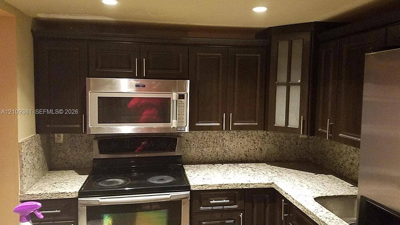 Kitchen featuring dark wood cabinetry, speckled stone countertops, built-in stainless steel microwave, recessed ceiling lighting, and glass-panel upper cabinet doors
