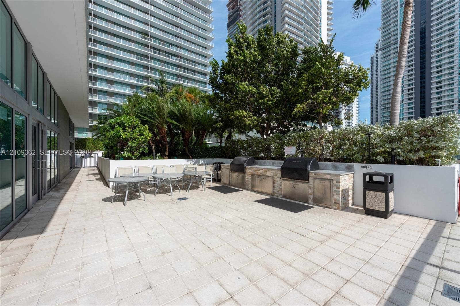 79 SW 12th Street, #1107-S, Miami, FL 33130 Outdoor terrace featuring light-toned tile flooring, built-in outdoor kitchen with stone veneer base, integrated stainless steel grills, and mature landscaping