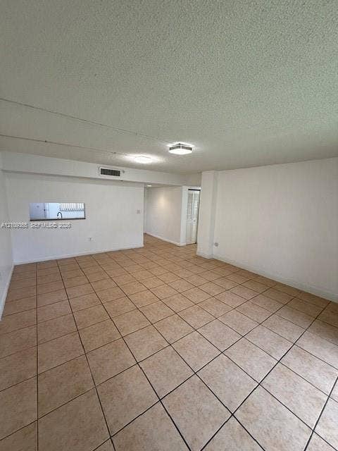 Open floor plan featuring neutral-tone ceramic tile flooring, textured ceilings, dual flush-mount light fixtures, and a wall cutout revealing a glimpse of kitchen fixtures