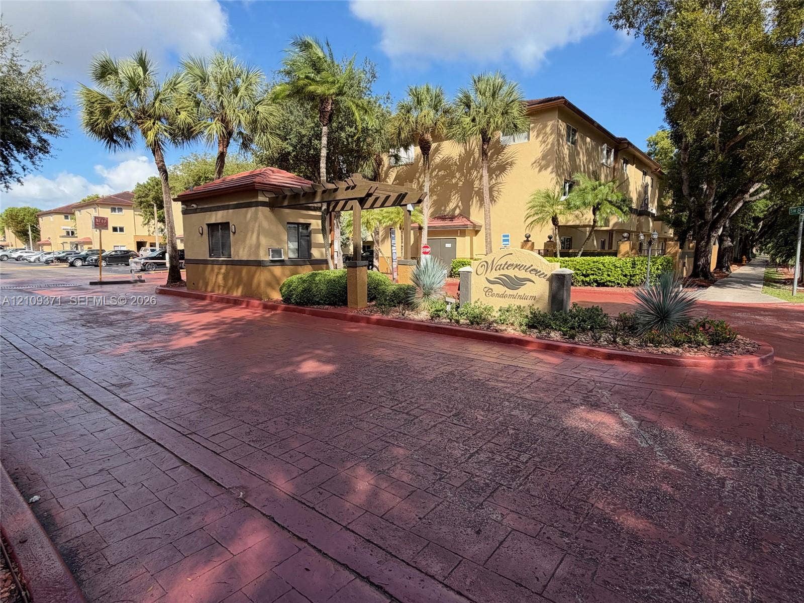 Gated entrance featuring a stamped concrete drive, an attended guard house, and mature palm landscaping