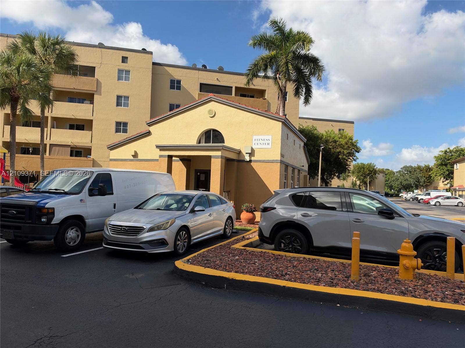 Stucco fitness center building featuring a gable roof, arched decorative window, and mature palm trees