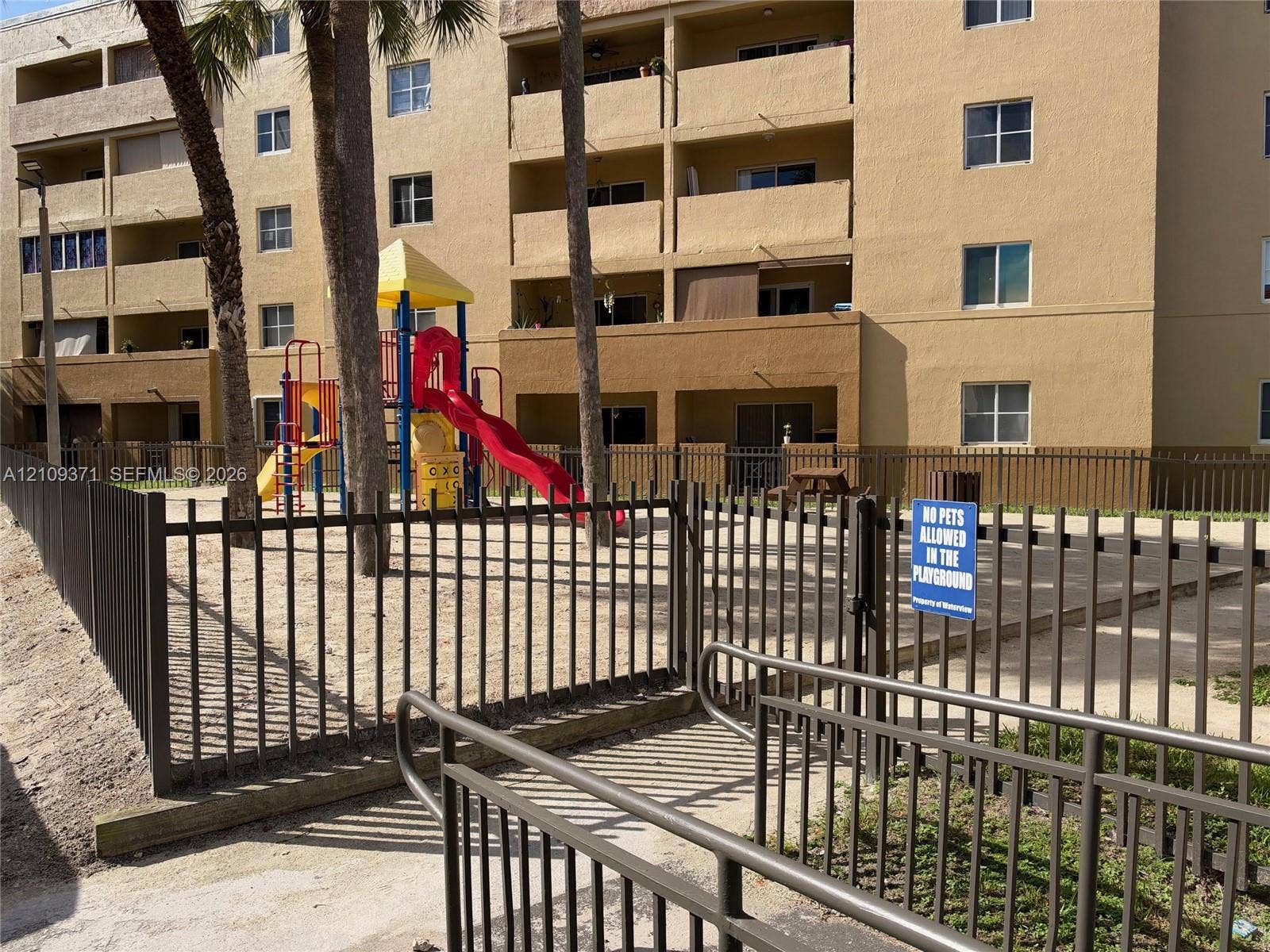 Fenced playground area featuring recreational equipment, sandy ground surface, and metal perimeter security fencing