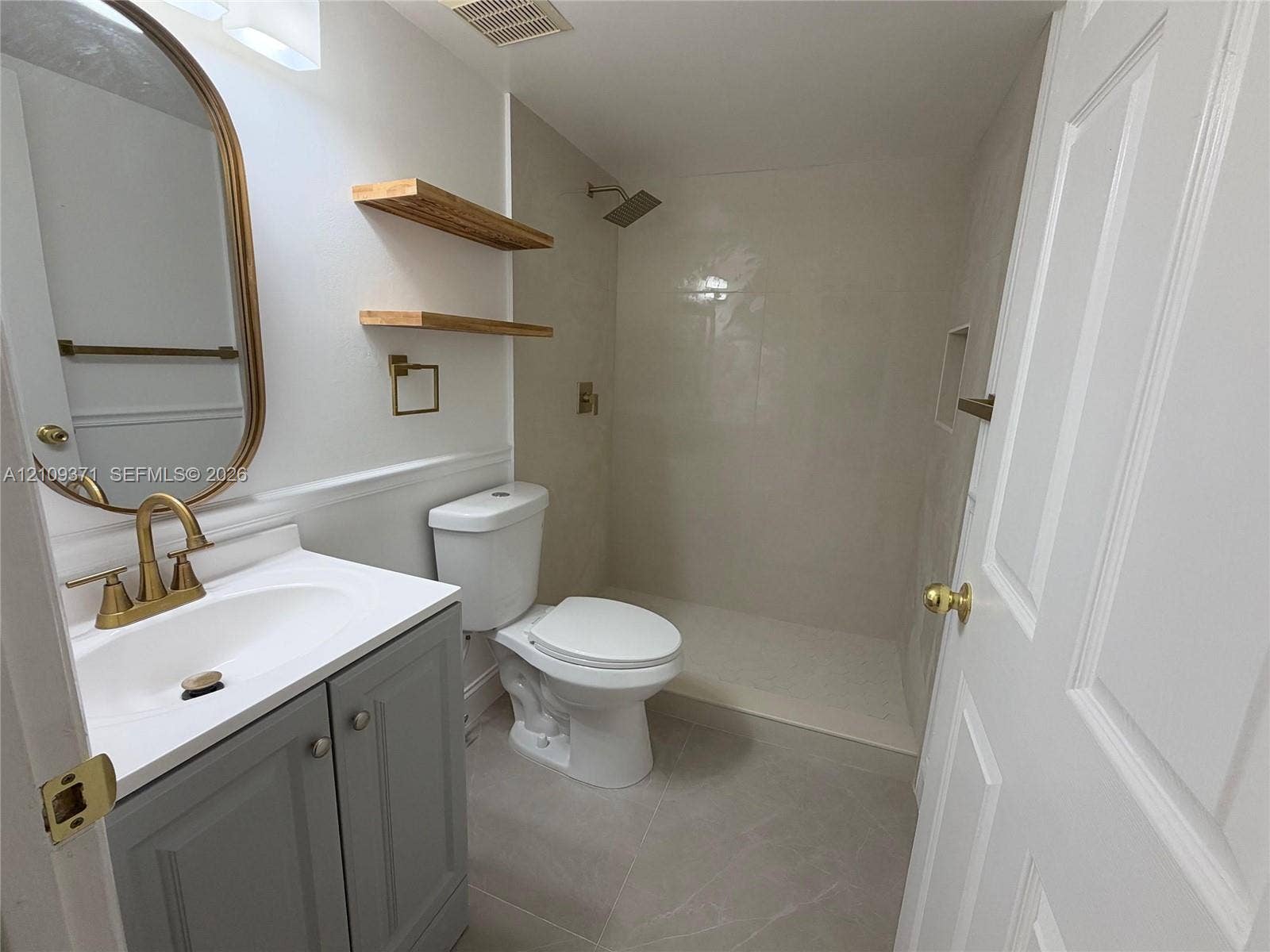 Bathroom featuring a grey vanity with a white countertop, brass-finish faucet and hardware, walk-in shower with large-format tile surround, and floating wood shelving
