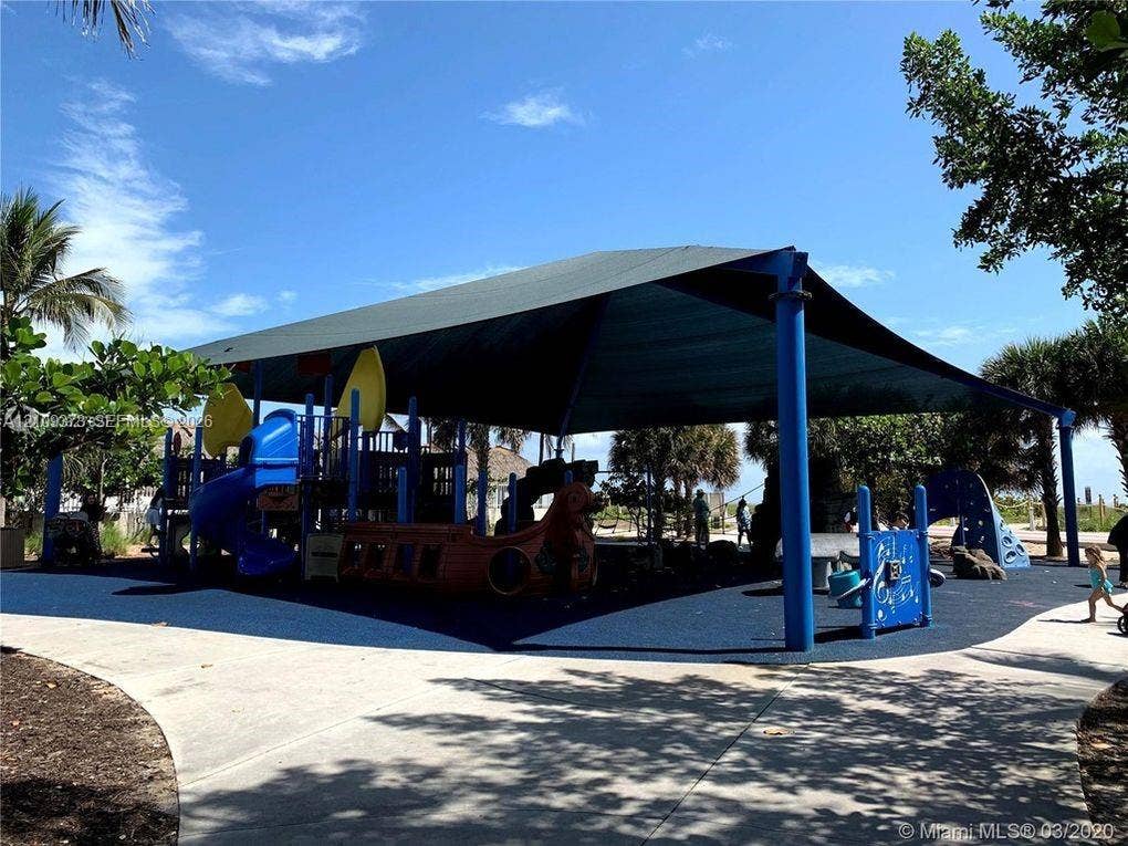 Recreational area featuring a large fabric shade structure, integrated play equipment, and a paved walkway surface