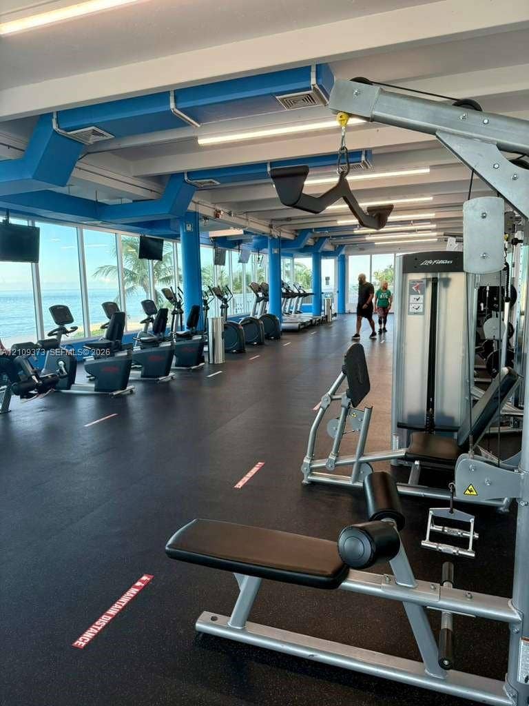 Fitness center featuring rubber flooring, exposed industrial-style blue ceiling ductwork, and floor-to-ceiling windows with coastal scenery